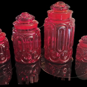May include: Four ruby red glass jars with lids. The jars vary in size and have a decorative pattern of circles and arches. The jars are displayed on a reflective surface against a black background.