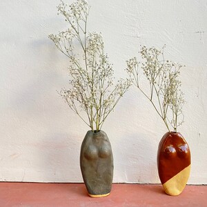 May include: Two ceramic vases in the shape of a human torso, one brown with a yellow bottom and the other gray with a gold bottom. Both vases are filled with white baby's breath flowers.