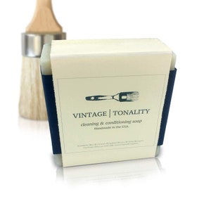 May include: A bar of soap with a white label that reads "VINTAGE | TONALITY cleaning & conditioning soap Handmade in the USA." The label also features a small illustration of a paintbrush. The soap is wrapped in a dark blue band.