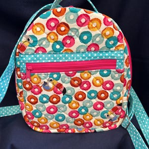 May include: A quilted backpack featuring a colorful pattern of toucans and circular shapes in shades of pink, orange, and teal. The bag includes a front zippered pocket with a bright pink zipper and light blue straps with white polka dots.