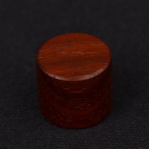 May include: A cylindrical, solid wood object with a rich, reddish-brown hue. The top and sides are smooth, and the object is set against a dark background. This could be a decorative element or a component of a larger item.