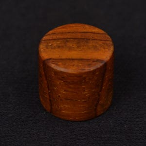 May include: A small, cylindrical wooden object with a flat top and bottom. The wood has a warm, reddish-brown hue with visible grain patterns. The object is likely a decorative element or a small stopper.
