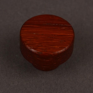 May include: A circular cabinet knob crafted from dark reddish-brown wood. The knob features a smooth, flat top surface with a visible horizontal wood grain pattern and a slightly tapered cylindrical base that connects to the mounting point.