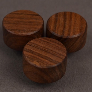Op de afbeelding: Drie ronde houten knoppen in een rijke bruine kleur met zichtbare natuurlijke houtnerven. Elke knop heeft een glad, plat bovenoppervlak en cilindrische zijkanten, wat de organische textuur en de diepe tinten van het houtmateriaal benadrukt.
