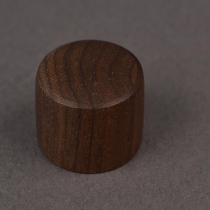 Op de afbeelding: Een cilindrisch object van donkerbruin hout met een platte boven- en onderkant. De houtnerf is zichtbaar. Het kan een flesstopper of een kleine decoratieve knop zijn. Het object is ongeveer 3 cm hoog.