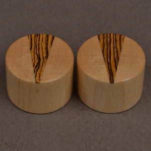 May include: Two cylindrical wooden plugs. Each has a light wood base and a V-shaped inlay of dark, striped wood. They are displayed on a neutral grey background, highlighting the natural wood grain and contrasting colours.