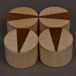 May include: Four small, cylindrical wooden boxes. Each has a light wood body and a dark brown inlaid geometric design on the lid. The boxes are arranged in a square formation against a neutral background.