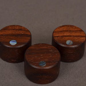 Peut inclure: Trois boutons en bois cylindriques avec un grain brun foncé. Chaque bouton comporte une petite incrustation ronde de matériau bleu et blanc irisé. Les boutons sont probablement faits à la main.