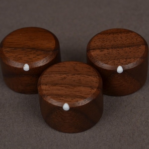 Peut inclure: Trois boutons en bois brun foncé, chacun avec un petit point blanc. Les boutons sont cylindriques et en bois. Ils sont disposés en triangle. Le grain du bois est visible sur les surfaces supérieures.