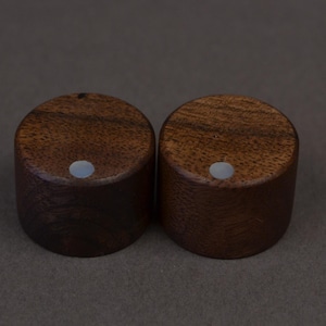 Könnte beinhalten: Zwei zylindrische Holzknöpfe mit dunkelbraunem Finish. Jeder Knopf hat einen kleinen, runden, hellfarbigen Einsatz auf der Oberseite. Die Knöpfe sind vor einem gedämpften braunen Hintergrund platziert, was eine minimalistische Ästhetik suggeriert.