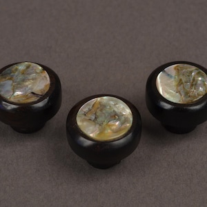 May include: Three dark brown cabinet knobs with a round, iridescent mother-of-pearl inlay. The knobs are set against a neutral gray background. These decorative knobs could be used for furniture or other home decor projects.