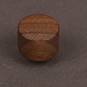 Op de afbeelding: Een kleine, cilindervormige houten knop met een gladde, gepolijste afwerking. Het object heeft een natuurlijk bruin houtnerfpatroon dat zichtbaar is op het platte bovenoppervlak en de gebogen zijkanten, wat de organische textuur benadrukt.