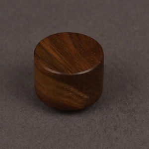 Op de afbeelding: Een klein, cilindrisch houten object met een afgeronde boven- en onderkant. De houtnerf is zichtbaar en toont een rijke bruine kleur. Het object staat tegen een neutrale grijze achtergrond.