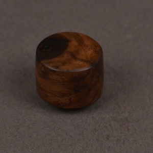 May include: A small, cylindrical wooden bottle stopper with a rounded top and a dark, circular pattern on the top. The stopper is a warm brown color and appears to be made of a polished wood.