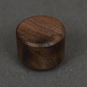 May include: A small, cylindrical wooden knob with a smooth, polished finish. The wood features a rich, dark brown color with visible natural grain patterns swirling across the flat top surface and the rounded sides of the object.