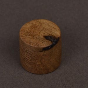 May include: A small, cylindrical wooden object with a flat top and bottom. The wood has a light brown color with darker grain patterns. A dark, irregular marking is visible on the top surface. The object is set against a dark gray background.