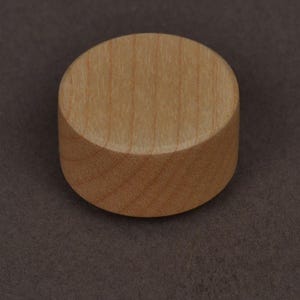 Op de afbeelding: Een klein, cilindrisch houten object met een platte boven- en onderkant. Het hout heeft een lichtbruine kleur en zichtbare nerf. Het object ligt op een donkerbruin oppervlak. Dit kan een klein meubelonderdeel of een decoratief element zijn.