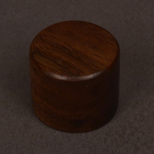 May include: A cylindrical, solid wood object with a flat top and bottom. The wood has a rich, dark brown color with visible grain patterns. The object is set against a dark brown background.