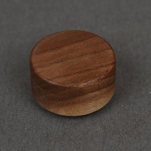 May include: A small, cylindrical wooden button with a smooth, polished finish. The wood features a natural medium-brown grain pattern with visible horizontal lines running across the flat, circular top surface of the button.