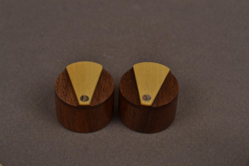 Pu&ograve; includere: Due manopole cilindriche realizzate in legno marrone scuro, ciascuna caratterizzata da un inserto triangolare in legno chiaro sulla superficie superiore. Un piccolo accento circolare iridescente simile a una conchiglia &egrave; incastonato.