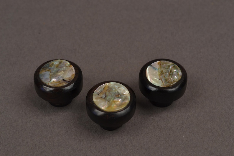 May include: Three dark brown cabinet knobs with a round, iridescent mother-of-pearl inlay. The knobs are set against a neutral gray background. These decorative knobs could be used for furniture or other home decor projects.