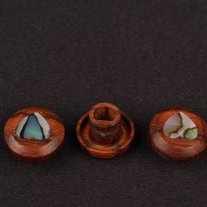 May include: Three wooden bottle stoppers with inlaid iridescent abalone shells. The stoppers are a rich brown colour and have a smooth, polished finish. One stopper is a cylinder, and the other two are round.