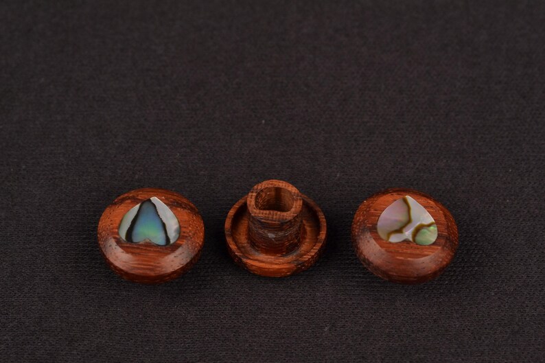 May include: Three wooden bottle stoppers with inlaid iridescent abalone shells. The stoppers are a rich brown colour and have a smooth, polished finish. One stopper is a cylinder, and the other two are round.