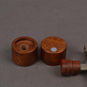 K&ouml;nnte beinhalten: Zwei kleine, zylindrische Holzknebel in einem satten, r&ouml;tlich-braunen Farbton. Einer der Kn&ouml;pfe hat ein zentrales Loch, w&auml;hrend der andere einen kleinen, grauen kreisf&ouml;rmigen Magneten hat. Ein Teil eines braunen Leder- und Metallbeschlags ist ebenfalls sichtbar.