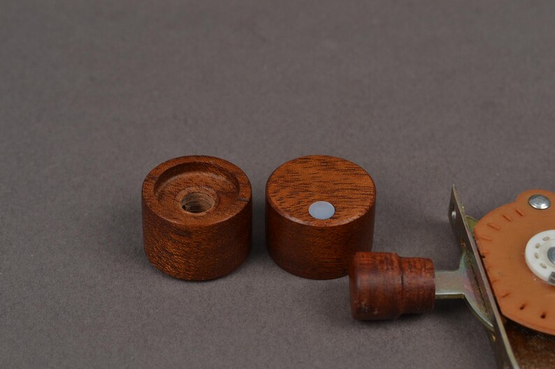 K&ouml;nnte beinhalten: Zwei kleine, zylindrische Holzknebel in einem satten, r&ouml;tlich-braunen Farbton. Einer der Kn&ouml;pfe hat ein zentrales Loch, w&auml;hrend der andere einen kleinen, grauen kreisf&ouml;rmigen Magneten hat. Ein Teil eines braunen Leder- und Metallbeschlags ist ebenfalls sichtbar.