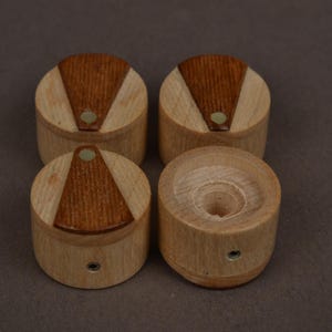 May include: A set of four cylindrical wooden knobs featuring a light-colored wood body with a darker, triangular wood inlay on the top surface. Each knob includes a small circular metal insert on the side for attachment and a central hole.