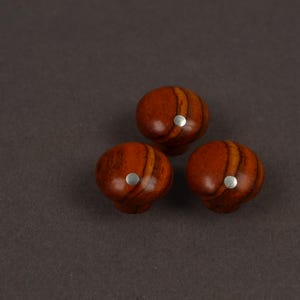 May include: Three round cabinet knobs made of polished wood with a distinct reddish-brown grain pattern. Each knob features a smooth, domed surface and a small, circular silver-colored metal inlay centered on the front face of the wooden knob.