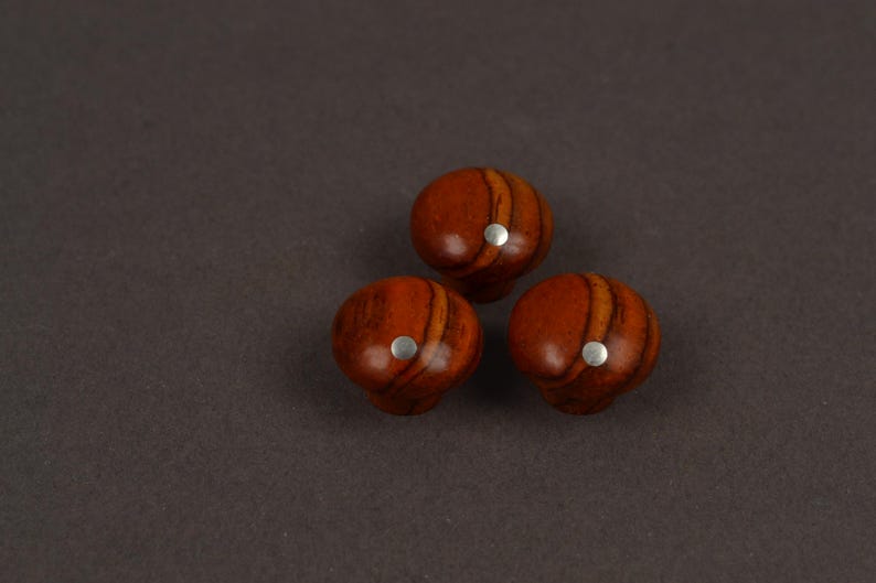 May include: Three round cabinet knobs made of polished wood with a distinct reddish-brown grain pattern. Each knob features a smooth, domed surface and a small, circular silver-colored metal inlay centered on the front face of the wooden knob.