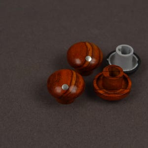 May include: Three round knobs with a polished reddish-brown wood grain finish. Two knobs are shown from the top, each featuring a small silver circular inlay. One knob is flipped over to reveal a grey plastic mounting base with a central slot.