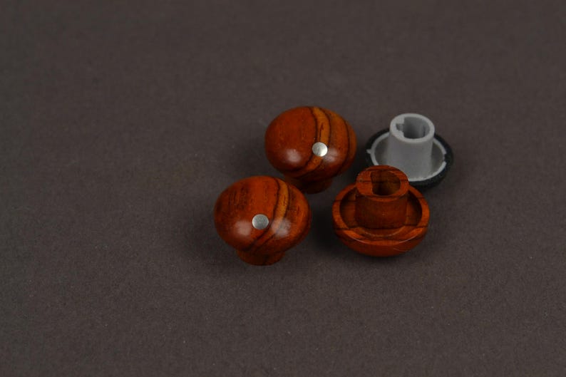 May include: Three round knobs with a polished reddish-brown wood grain finish. Two knobs are shown from the top, each featuring a small silver circular inlay. One knob is flipped over to reveal a grey plastic mounting base with a central slot.
