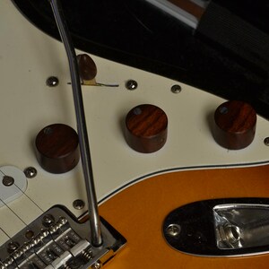 May include: Close-up of an electric guitar with a cream-colored pickguard and a sunburst finish. The guitar features three wooden control knobs, a chrome tremolo arm, and six strings. The body has a gradient of orange, yellow, and brown.