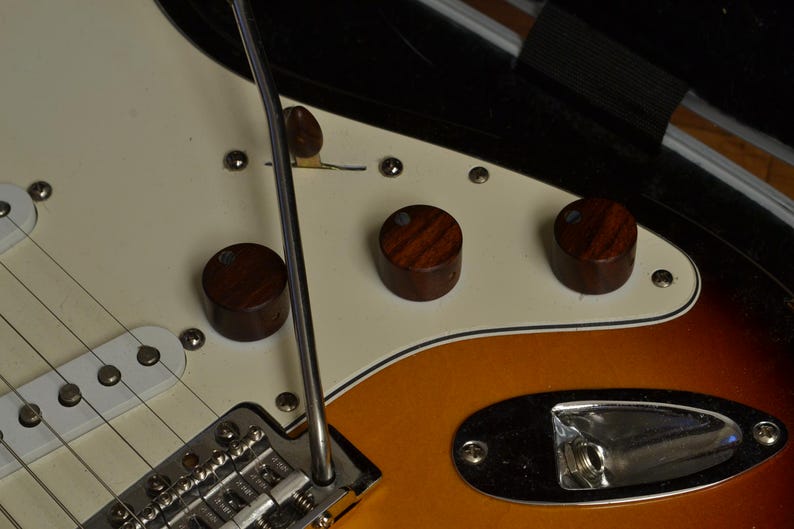 May include: Close-up of an electric guitar with a cream-colored pickguard and a sunburst finish. The guitar features three wooden control knobs, a chrome tremolo arm, and six strings. The body has a gradient of orange, yellow, and brown.