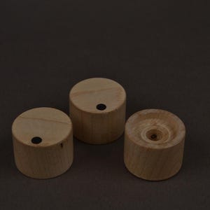 May include: Three small, cylindrical wooden objects with a light brown color. Each has a flat top and a dark dot. One has a central hole. The objects are arranged on a dark brown surface.