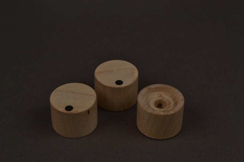 May include: Three small, cylindrical wooden objects with a light brown color. Each has a flat top and a dark dot. One has a central hole. The objects are arranged on a dark brown surface.