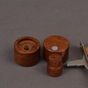 K&ouml;nnte beinhalten: Drei Holzkn&ouml;pfe in verschiedenen Gr&ouml;&szlig;en mit einer satten braunen Oberfl&auml;che. Zwei sind zylindrisch, einer ist k&uuml;rzer und runder. Eine Metall- und Lederkomponente ist teilweise rechts sichtbar, was auf ein M&ouml;bel- oder Hardware-Thema hindeutet.