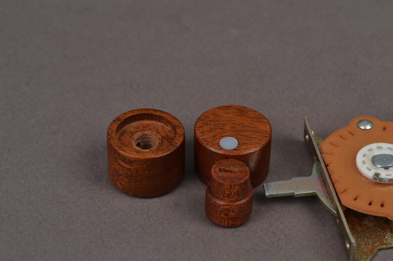 K&ouml;nnte beinhalten: Drei Holzkn&ouml;pfe in verschiedenen Gr&ouml;&szlig;en mit einer satten braunen Oberfl&auml;che. Zwei sind zylindrisch, einer ist k&uuml;rzer und runder. Eine Metall- und Lederkomponente ist teilweise rechts sichtbar, was auf ein M&ouml;bel- oder Hardware-Thema hindeutet.