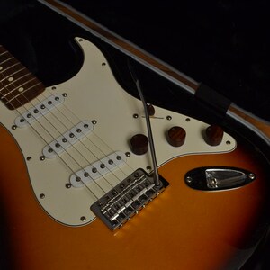 May include: A sunburst-finish electric guitar with a white pickguard and chrome hardware. The guitar has three brown control knobs and a tremolo bar. The instrument is in an open black case, ready to play.