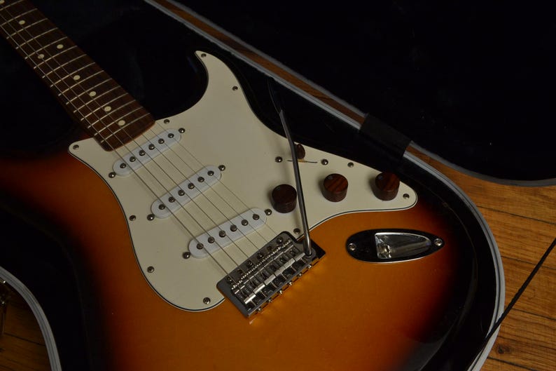 May include: A sunburst-finish electric guitar with a white pickguard and chrome hardware. The guitar has three brown control knobs and a tremolo bar. The instrument is in an open black case, ready to play.