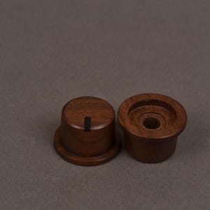 May include: Two cylindrical dark brown wooden knobs with a smooth finish. One knob is positioned upright showing a flat top with a single vertical black indicator line, while the other is inverted to reveal a hollow center with a circular opening.