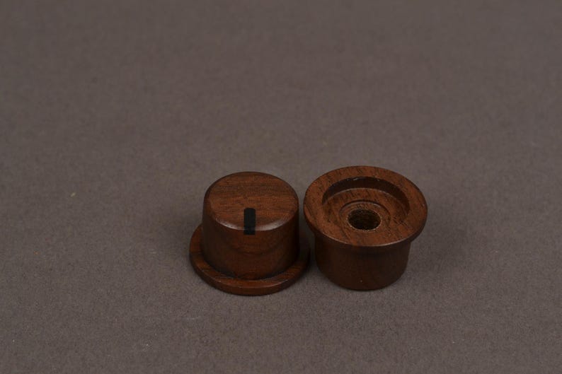May include: Two cylindrical dark brown wooden knobs with a smooth finish. One knob is positioned upright showing a flat top with a single vertical black indicator line, while the other is inverted to reveal a hollow center with a circular opening.