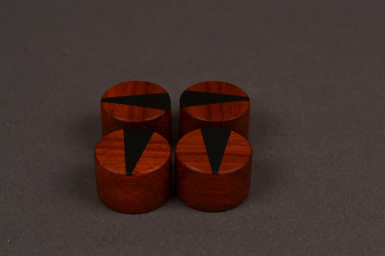 Peut inclure: Quatre objets cylindriques en bois de couleur brun rouge&acirc;tre. Chacun pr&eacute;sente une incrustation noire, triangulaire ou trap&eacute;zo&iuml;dale sur la surface sup&eacute;rieure. Ils sont dispos&eacute;s en carr&eacute; sur un fond neutre.