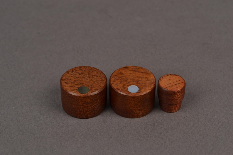 K&ouml;nnte beinhalten: Drei Holzkn&ouml;pfe in verschiedenen Gr&ouml;&szlig;en. Die Kn&ouml;pfe sind zylindrisch mit einer dunkelbraunen Oberfl&auml;che und einem kleinen, kreisf&ouml;rmigen Einsatz. Der kleinste Knopf hat eine abgerundete Oberseite, w&auml;hrend die gr&ouml;&szlig;eren flache Oberseiten haben. Sie sind wahrscheinlich f&uuml;r M&ouml;bel oder Schr&auml;nke.