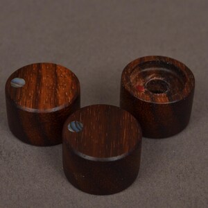 May include: Three dark brown wooden knobs with a cylindrical shape. Each knob has a circular, inlaid iridescent shell accent. One knob has a hole in the center. The knobs are arranged on a neutral gray background.