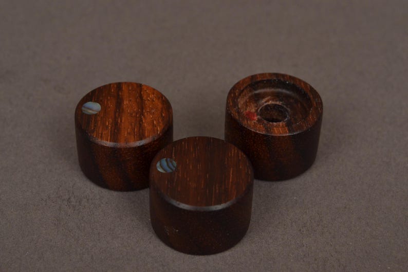 May include: Three dark brown wooden knobs with a cylindrical shape. Each knob has a circular, inlaid iridescent shell accent. One knob has a hole in the center. The knobs are arranged on a neutral gray background.