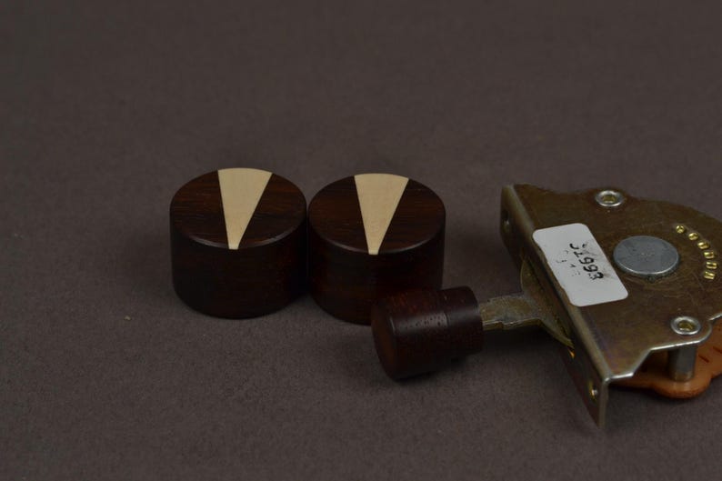 Puede incluir: Dos perillas cil&iacute;ndricas de madera de color marr&oacute;n oscuro con una incrustaci&oacute;n triangular de color claro en la parte superior, colocadas junto a un componente mec&aacute;nico de metal con una perilla de madera m&aacute;s peque&ntilde;a y una etiqueta blanca con el texto 31993.