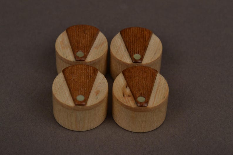 May include: Four cylindrical wooden knobs arranged in a square formation. Each knob features a light-colored natural wood base with a darker, triangular-shaped wood inlay on the top surface, accented by a small, circular, light-colored center dot.